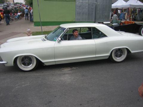 1996 Ford Bronco - A man drives a classic white Buick Riviera down a street, with a flea market in the background.