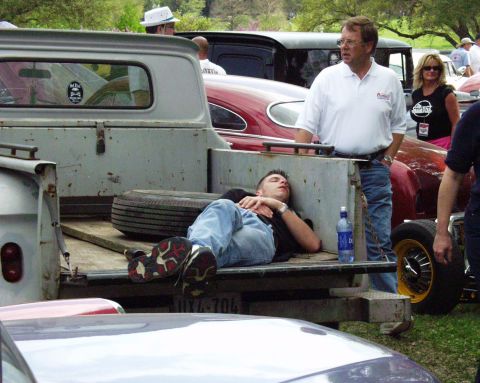 1996 Ford Bronco - A man rests in the bed of a pickup truck at a car show, surrounded by other vehicles and attendees.