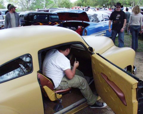 1996 Ford Bronco - A man is seated inside a customized hot rod, with the door open. Other vehicles and people are in the background.