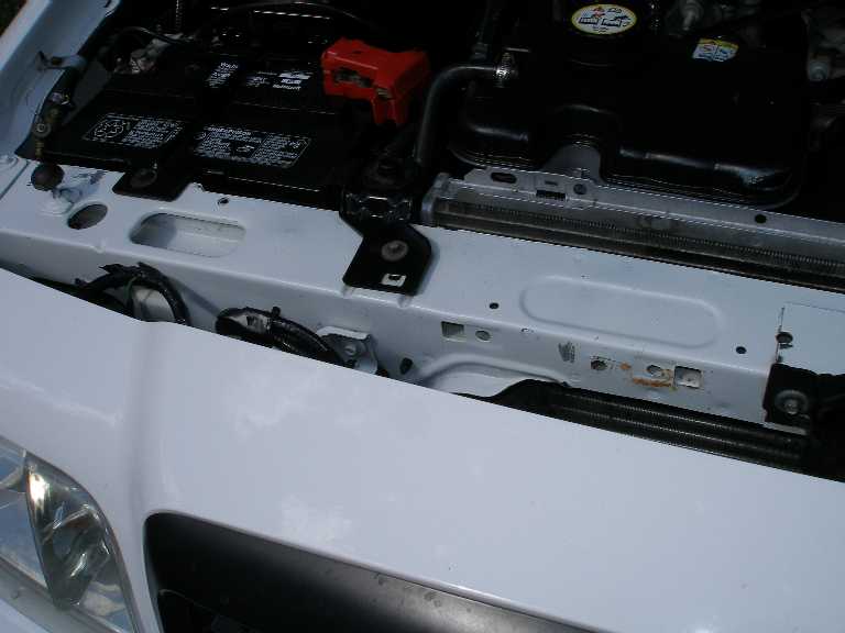 Engine bay of white car showing battery with red terminal cover and painted radiator core support