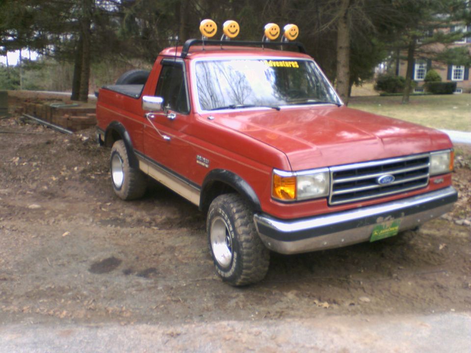 1989 Ford Bronco with smiley face lights on roof rack, parked on dirt