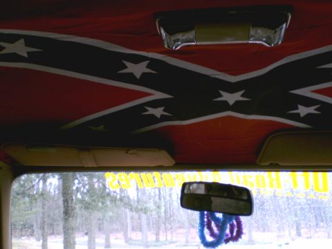 1989 Ford Bronco - The interior of a vehicle features a Confederate flag headliner and a rearview mirror adorned with fuzzy decorations.