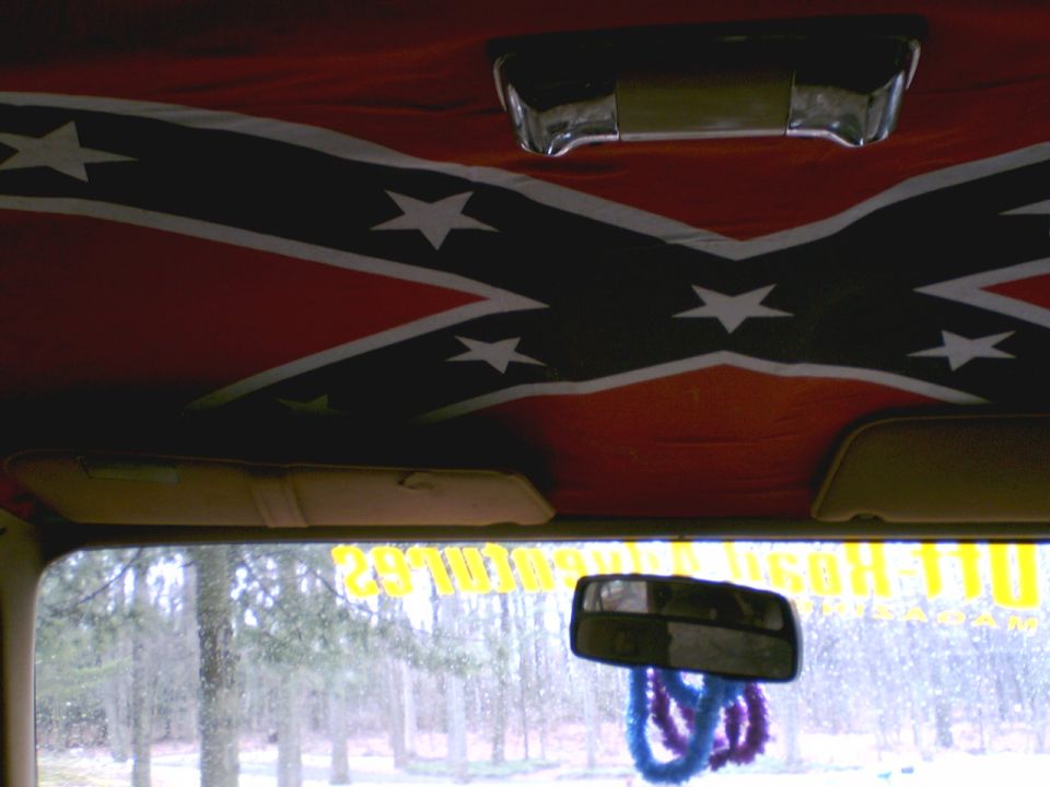 Inside a vehicle, looking up at a Confederate flag headliner. Sun visors are down. Rearview mirror hangs with blue and purple fuzzy things. Trees visible through the windshield