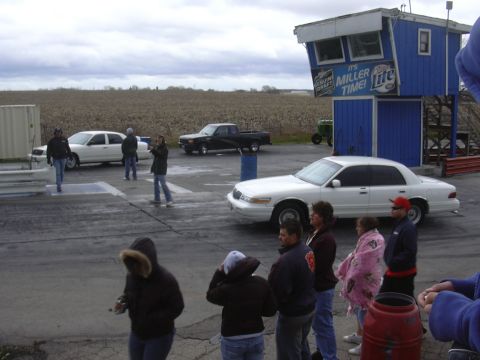 1996 Ford Crown Victoria - A group of people stand near a white sedan and a black pickup truck at a drag strip under a cloudy sky.
