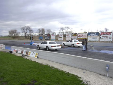 1996 Ford Crown Victoria - Two white sedans are lined up on a drag strip under an overcast sky, with various sponsor signs visible in the background.