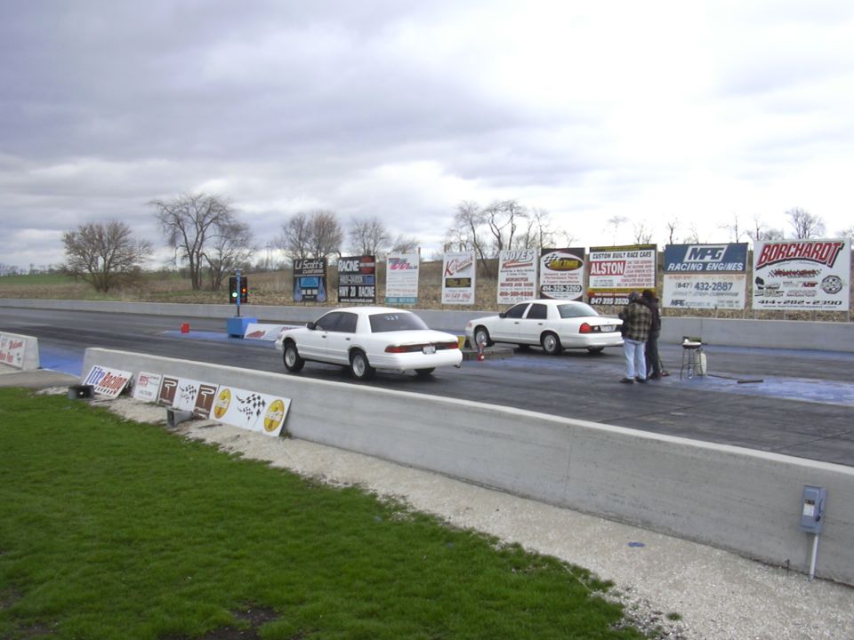 Two white 1996 Ford Crown Victorias on a drag strip, one in the foreground with its rear facing the camera, the other further down the track