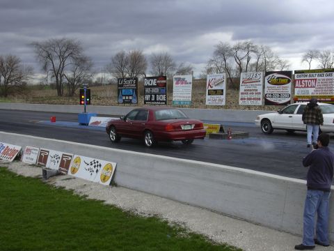 1996 Ford Crown Victoria - Two cars are lined up at a drag strip, ready for a race under a cloudy sky.