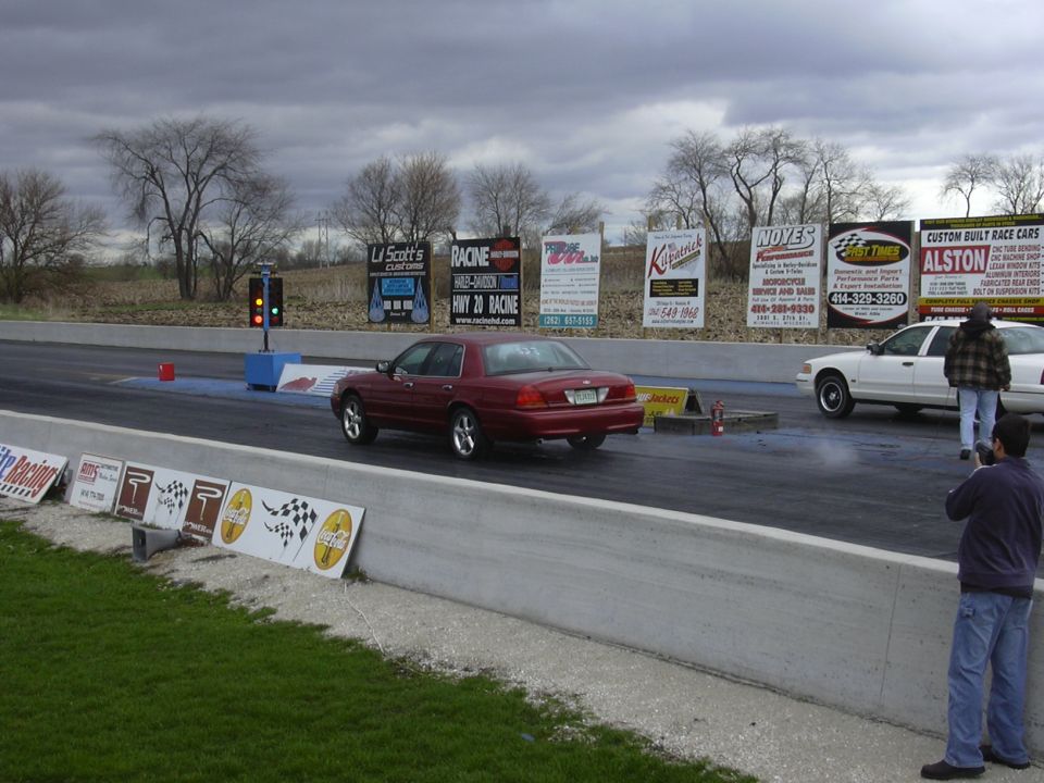 Red car and white car at drag strip starting line. Signs in background