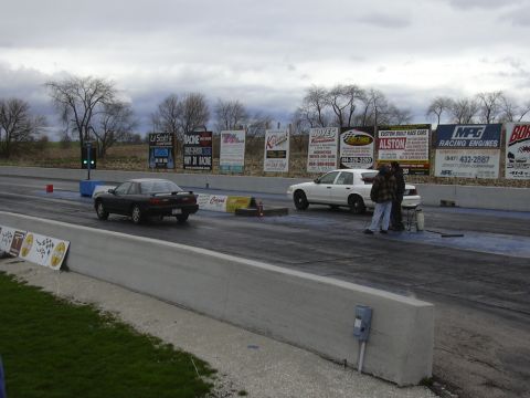 1996 Ford Crown Victoria - Two cars prepare for a drag race under an overcast sky, with numerous sponsor signs visible in the background.