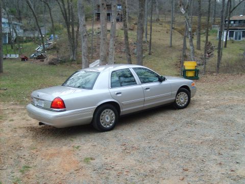 2004 Ford Crown Victoria - A silver sedan is parked outdoors on a gravel surface, with trees and houses in the background.