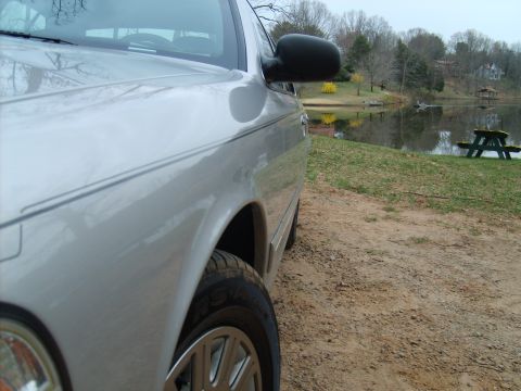 2004 Ford Crown Victoria - A silver 2004 Ford Crown Victoria is parked on a dirt path next to a lake with a picnic table in the background.