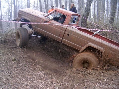 1979 Jeep J-10 - A lifted pickup truck navigates a muddy, wooded terrain, with onlookers observing the challenging off-road course.