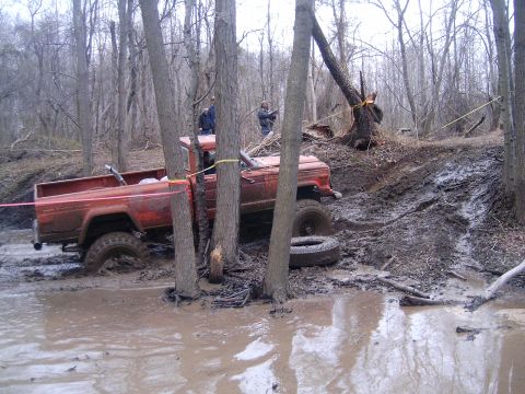 1979 Jeep J-10 - A red pickup truck navigates a muddy off-road course, with trees and debris visible.