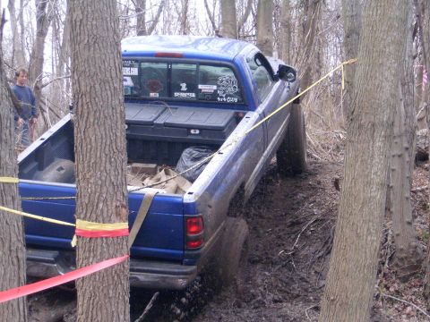 1979 Jeep J-10 - A blue pickup truck is stuck in the mud while navigating a wooded trail, with a person watching in the background.