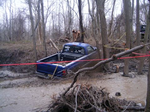 1979 Jeep J-10 - A blue pickup truck is stuck in the mud and trees, with another vehicle visible in the distance.