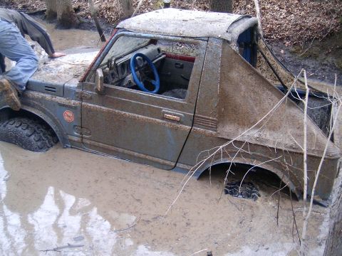 1979 Jeep J-10 - A mud-covered Suzuki Samurai is stuck in a muddy ditch, with a person attempting to get out.
