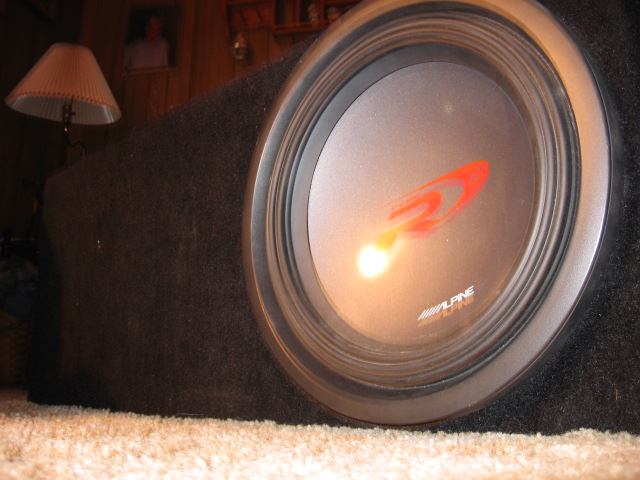 Car subwoofer in a box, showing the Alpine logo on the speaker cone. Carpeted floor visible