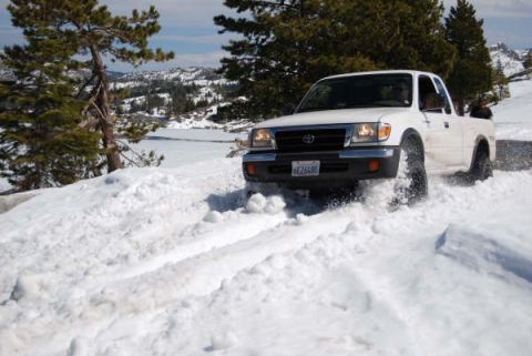 1989 Ford Bronco - A white Toyota Tacoma navigates a snowy mountain road, kicking up snow as it drives.