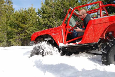 1989 Ford Bronco - A man drives a red off-road vehicle through a snowy landscape with pine trees in the background.