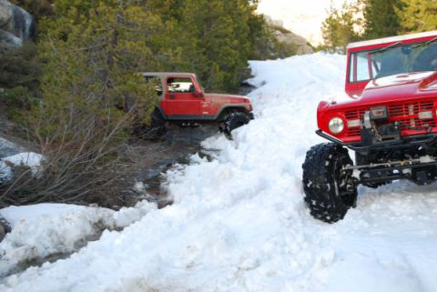 1989 Ford Bronco - Two off-road vehicles navigate a snowy landscape, one climbing a snowbank while the other is in a ditch.