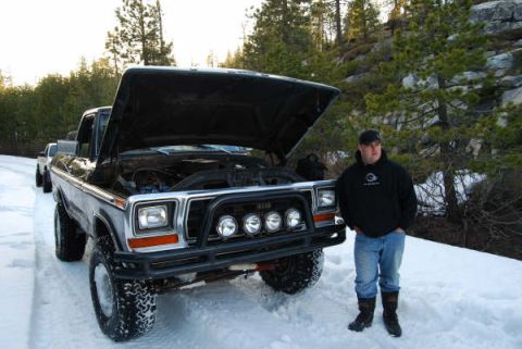 1989 Ford Bronco - A man stands by a lifted Ford F-Series truck with its hood up, parked on a snowy road with trees in the background.