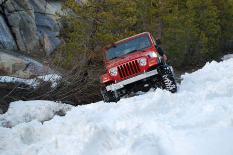 1989 Ford Bronco - A red Jeep Wrangler navigates a steep, snow-covered incline, with rocky terrain and pine trees visible behind it.