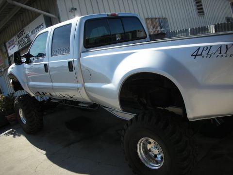 1998 Ford Crown Victoria - A silver pickup truck with large off-road tires and "4PLAY OFF ROAD" graphics is parked outside a business.