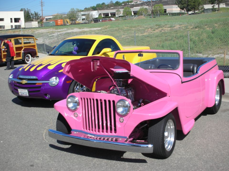 Pink open-top hot rod with raised hood beside a purple flamed Chevy SSR at a car show