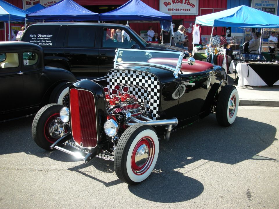Black hot rod roadster with checkered firewall, chrome engine, and whitewall tires at a show