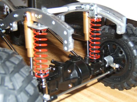 1988 Ford Bronco - Close-up of a scale RC rock-crawler chassis on a wood surface showing dual red coilover shocks, a black solid axle and knobby tires.