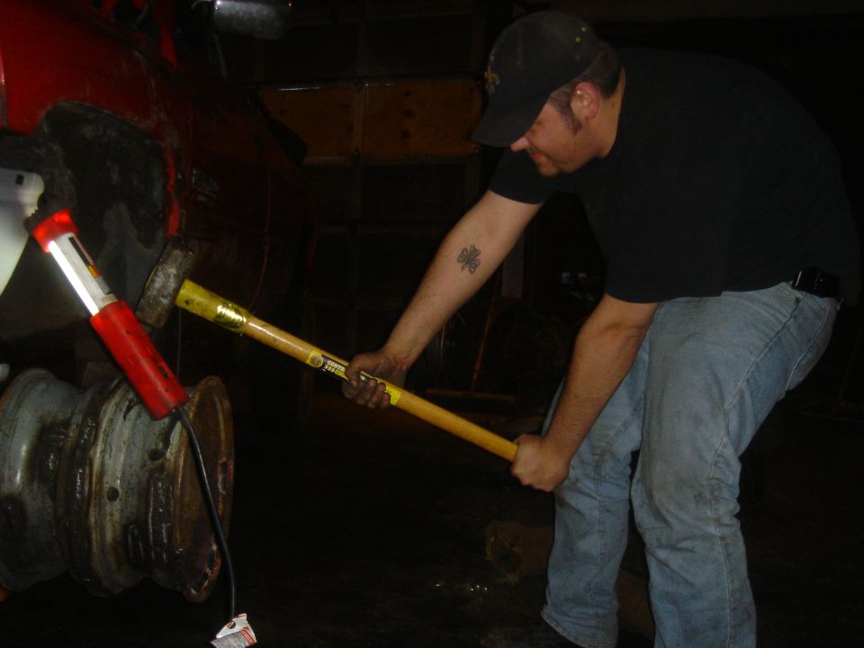 Man with sledgehammer working on a rusty vehicle part, illuminated by a red work light