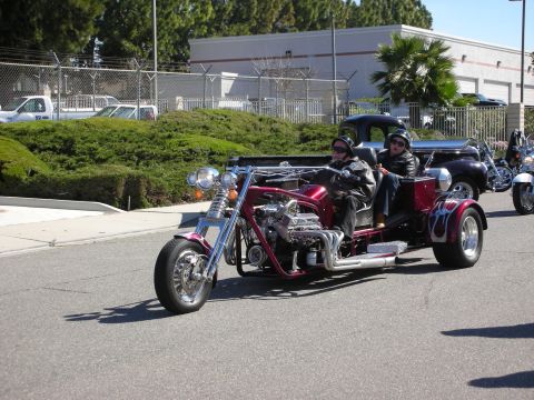 1998 Ford Crown Victoria - A custom-built trike with a large V8 engine and two occupants cruises down the road, with other vehicles visible in the background.