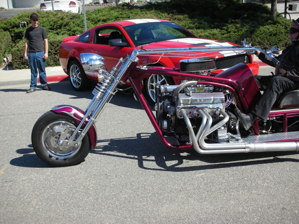 Purple custom V8 trike with long chrome forks beside a red Mustang at a car show