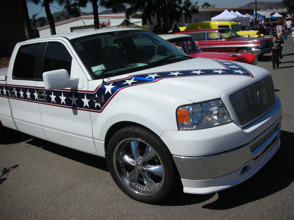 White Ford F-150 truck with stars-and-stripes graphics and chrome wheels at a car show