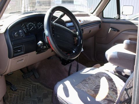 1996 Ford Bronco - Interior of a tan-and-grey full-size truck cab shot from the passenger door, showing the black dash, analog gauge cluster, aftermarket head…