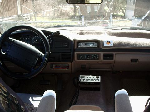 1996 Ford Bronco - Interior shot from the rear seat of a truck cab looking forward over a tan dashboard and black Ford steering wheel, with an aftermarket CD…
