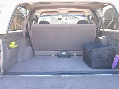 1996 Ford Bronco - Cargo bay of a tan-interior full-size SUV photographed through the open rear hatch, with the rear bench upright, tan carpet, and the factor…