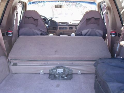 1996 Ford Bronco - View forward from the cargo area of a tan-interior full-size SUV with the rear bench folded down flat and the factory bottle jack strapped…