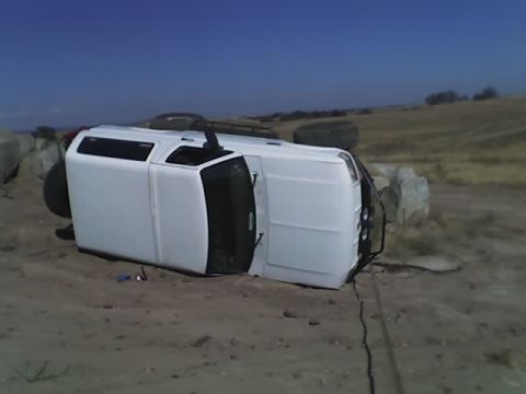 1995 Ford Bronco - A white SUV has rolled onto its side in a dry, rocky terrain under a clear blue sky.