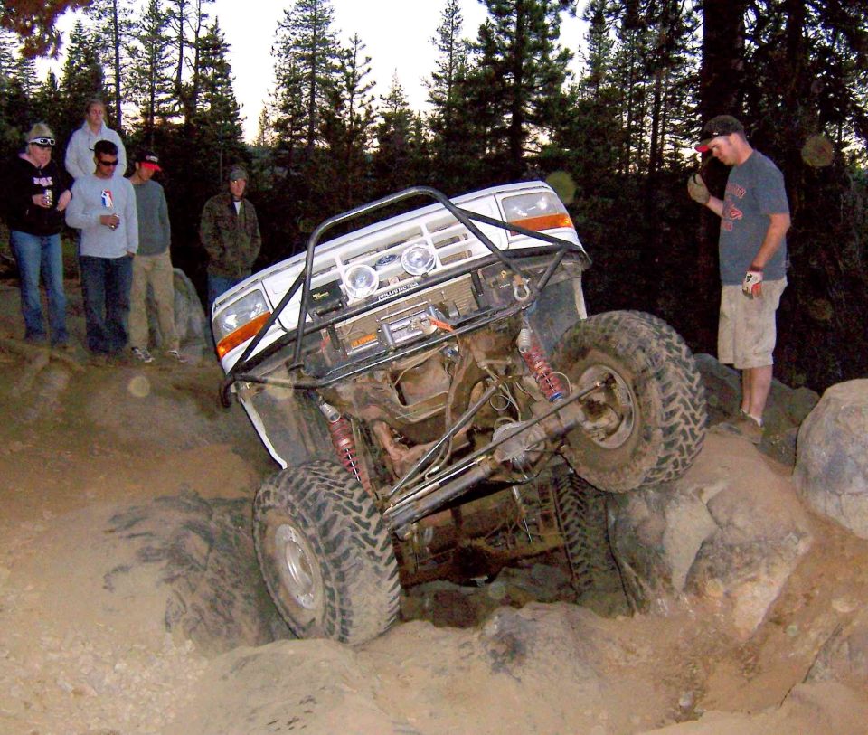 White Ford rock crawler SUV climbing over boulders showing undercarriage and big tires, with spectators