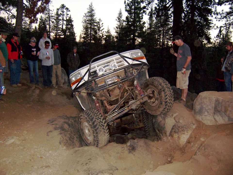 White SUV with roll cage going over rocks, people watching