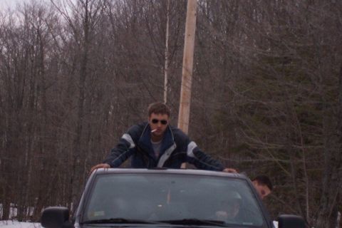 2000 Chevrolet Tahoe - A man stands on the roof of a vehicle, smoking a cigarette, with a wooded background.