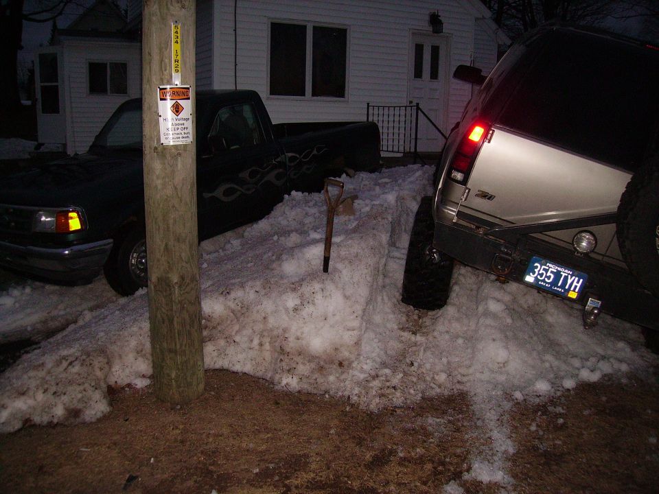 Rear view of a silver 1995 Chevrolet Tahoe with a license plate reading 355 TYH, stuck in snow next to a black pickup truck with flame decals