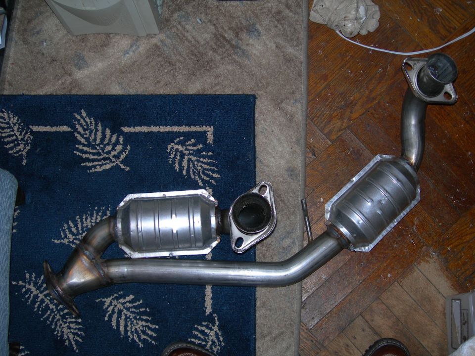 Exhaust pipe with two catalytic converter sections. One section is on a blue rug, the other is on a wood floor