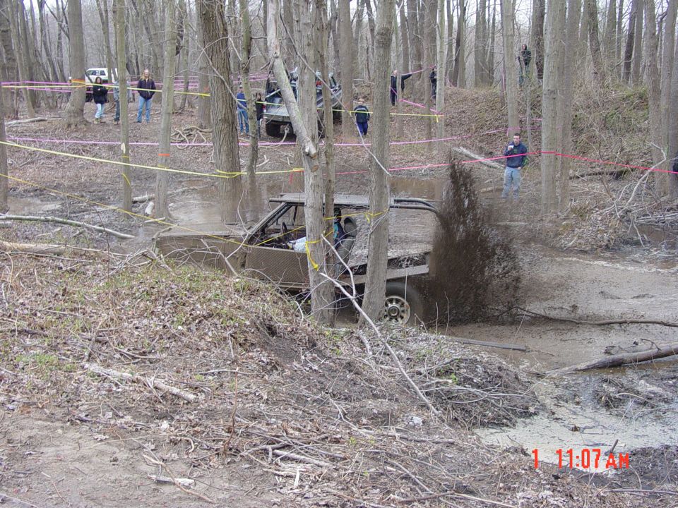 Mud-covered off-road truck with open doors driving through a muddy ditch, splashing water