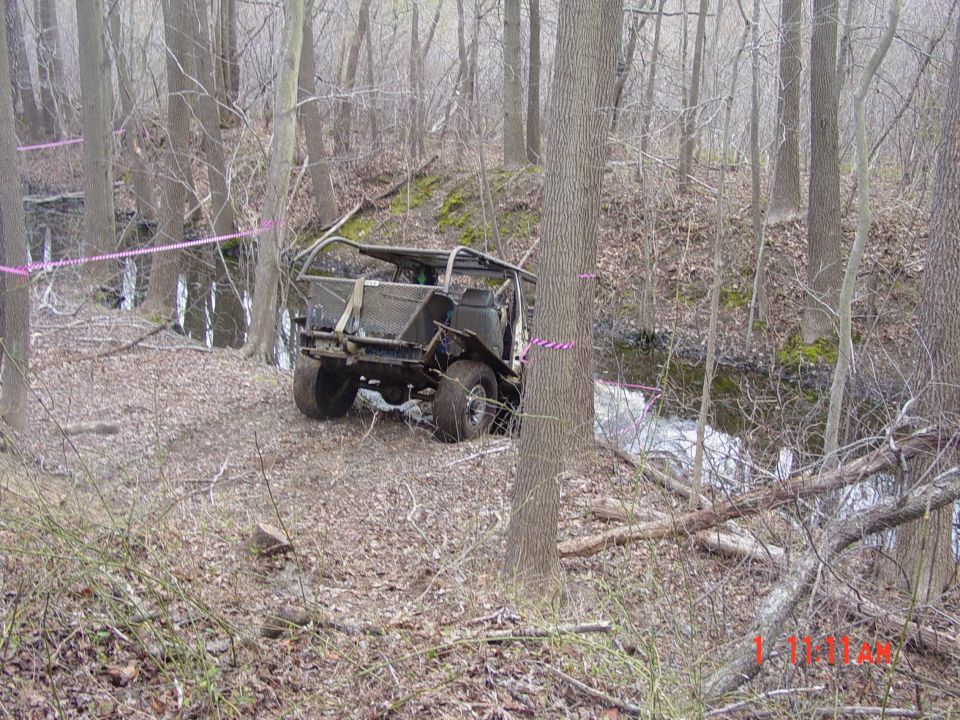 Off-road vehicle with no top or doors, parked on a wooded trail near a stream