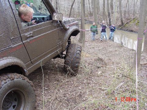 1995 Dodge 1500 - A muddy Suzuki Samurai navigates a wooded trail next to a shallow stream, with onlookers present.