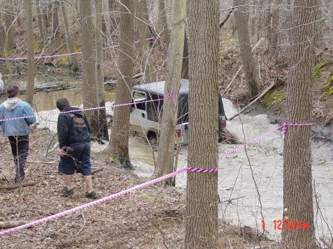 1995 Dodge 1500 - A silver Jeep Wrangler navigates a muddy, wooded obstacle course, observed by two individuals.