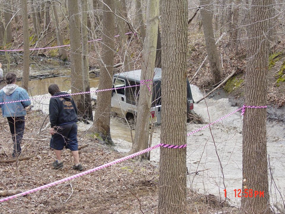 Silver Jeep Wrangler stuck in mud on a wooded trail, with two people observing