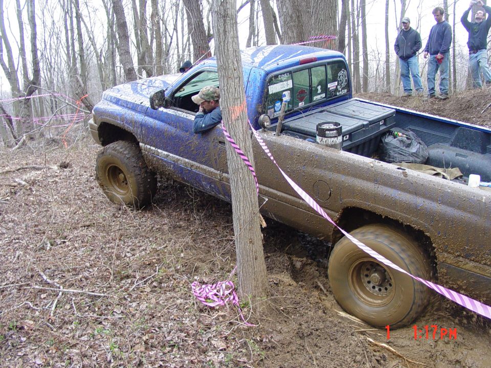 Blue pickup truck stuck in mud on a wooded trail, driver looking out window. Pink ribbon marks course